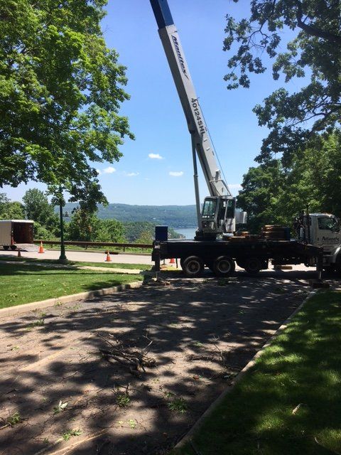 Crane on a truck near a road, lifting something; trees, hills, and blue sky in the background.