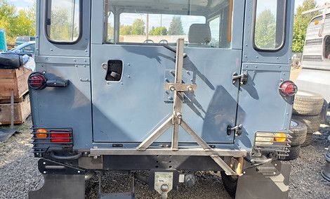 Rear view of a gray off-road vehicle with a rear-mounted tripod rack, parked outdoors on gravel.