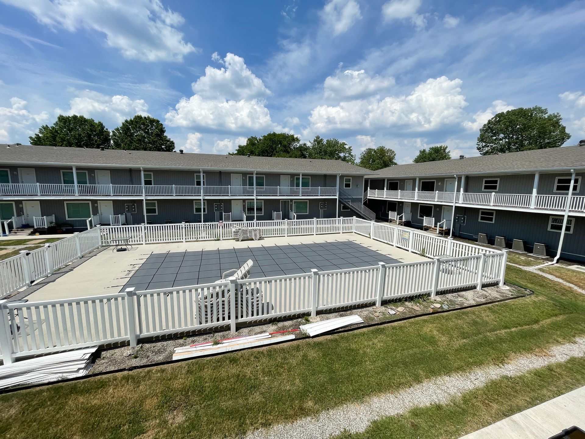Empty swimming pool surrounded by motel rooms and white fencing under a cloudy blue sky