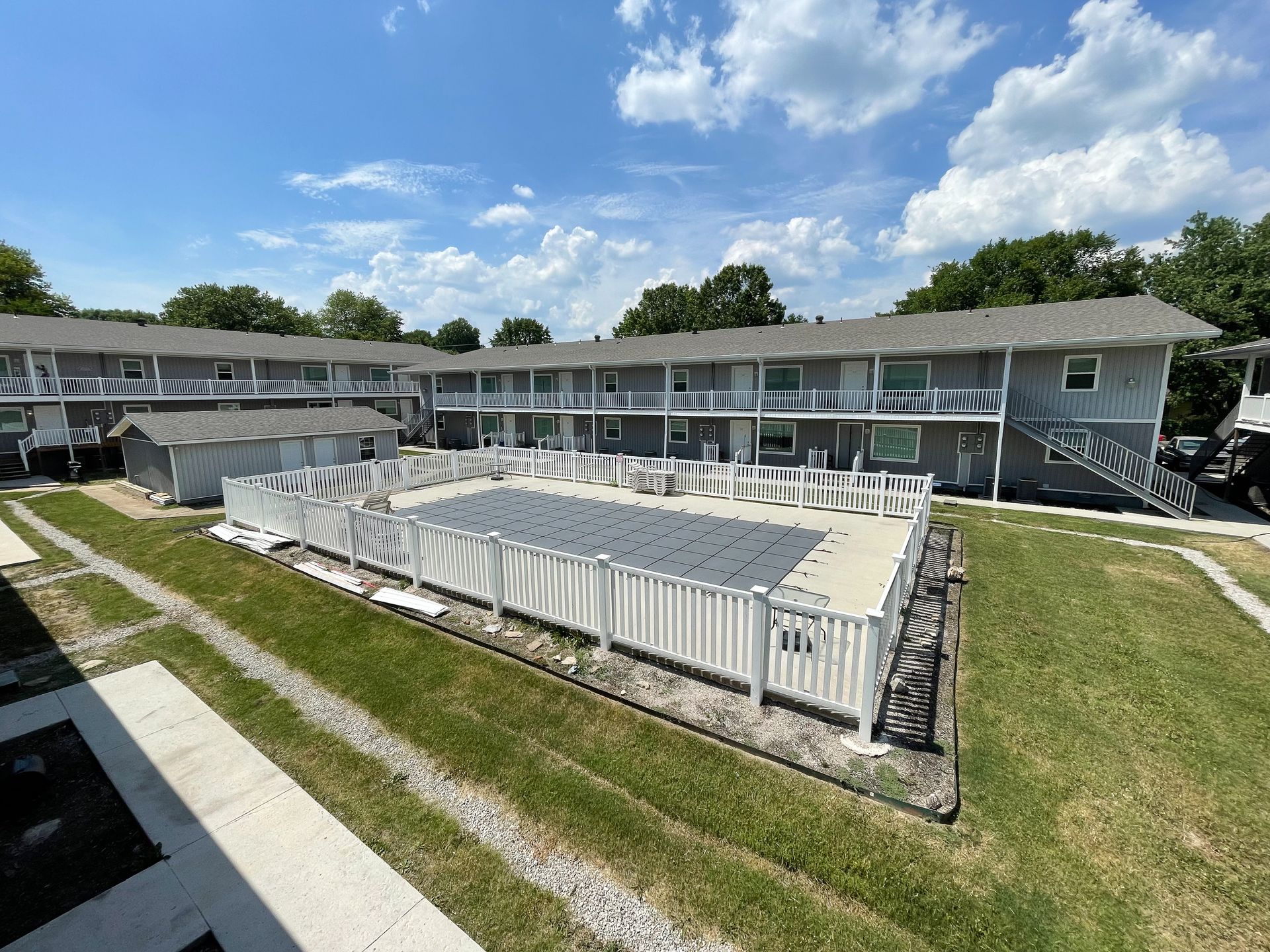 Apartment courtyard with a white fenced pool, green lawns, and two-story buildings under a blue sky