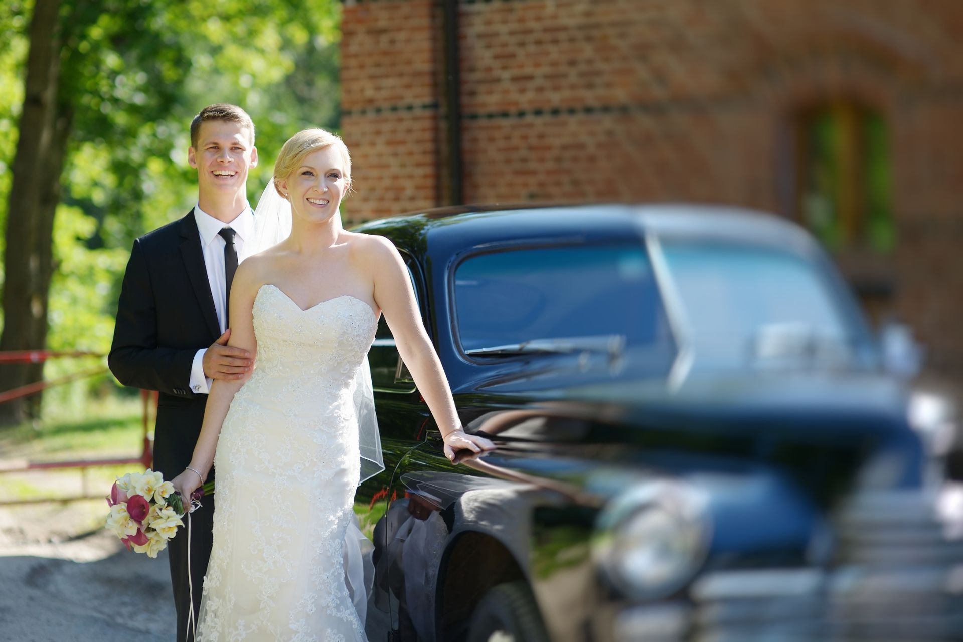 A bride and groom are posing for a picture in front of a black car.