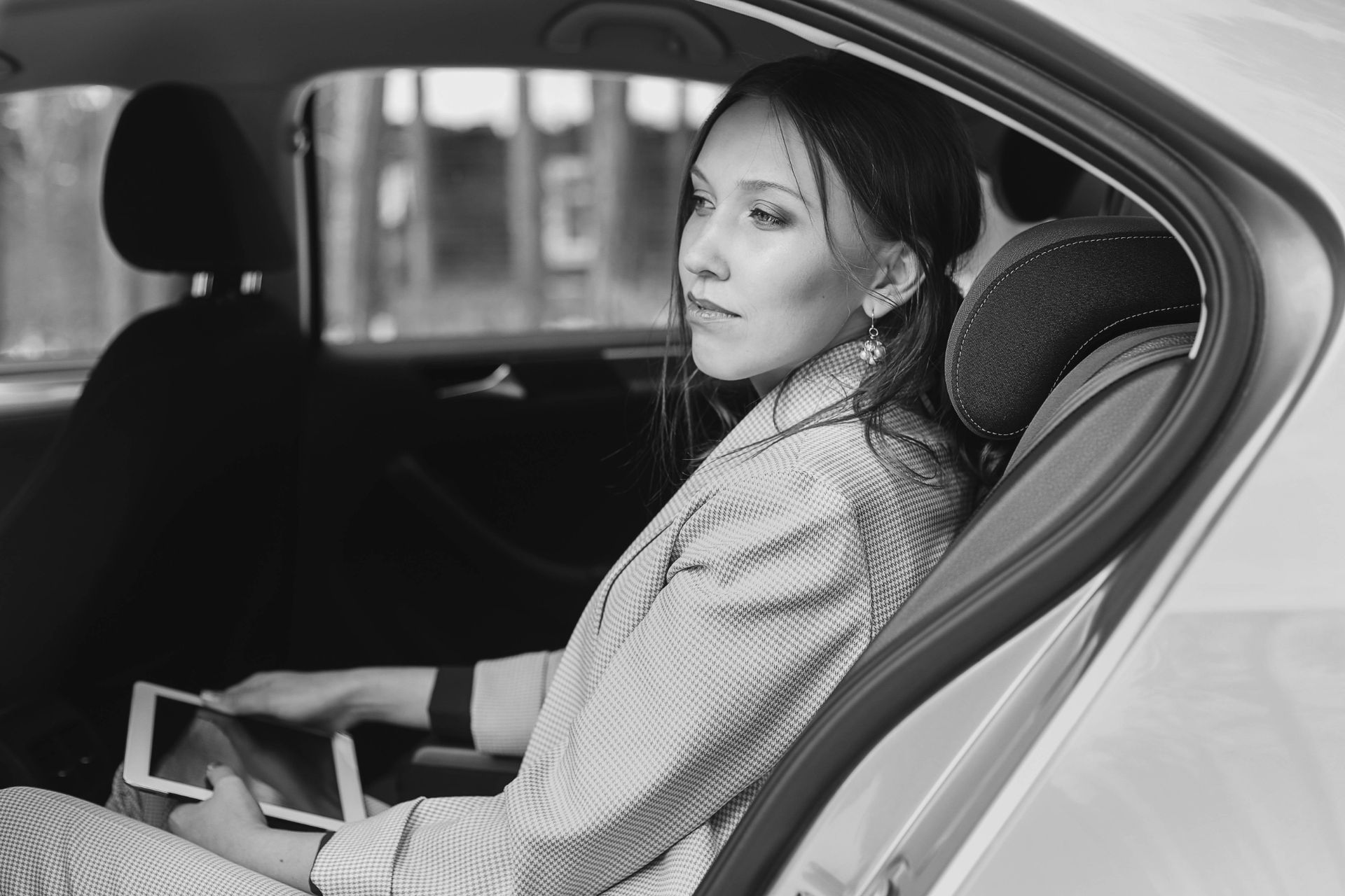 A woman is sitting in the back seat of a car using a tablet.