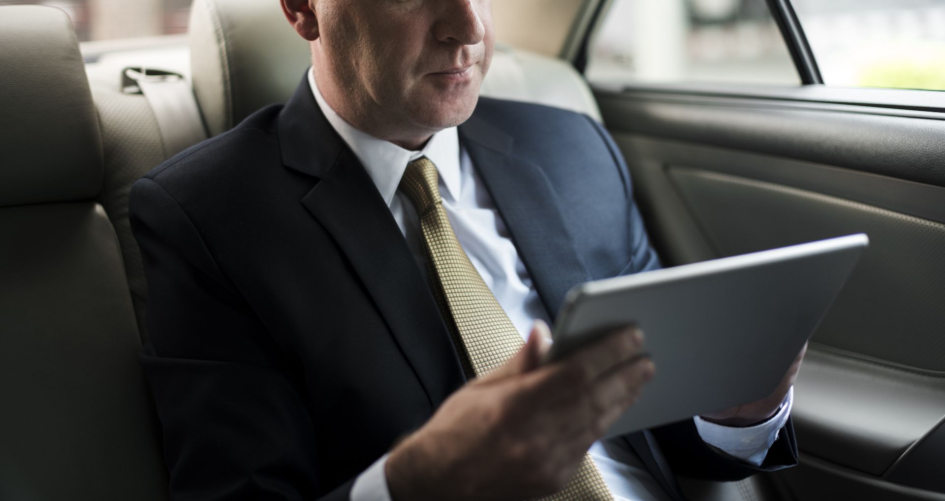 A man in a suit and tie is sitting in the back seat of a car using a tablet.