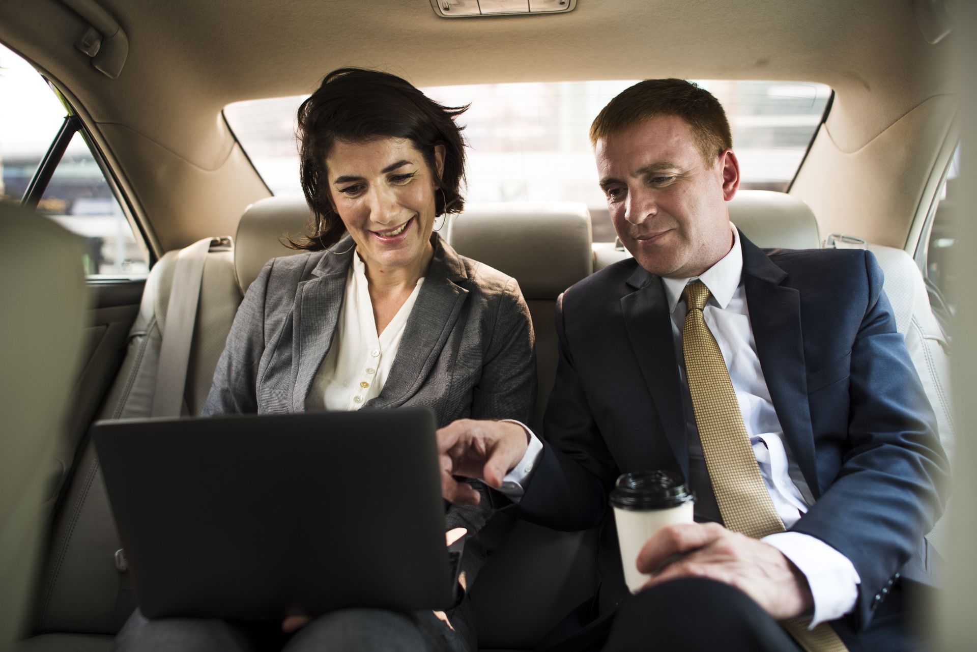 A man and a woman are sitting in the back seat of a car looking at a laptop.