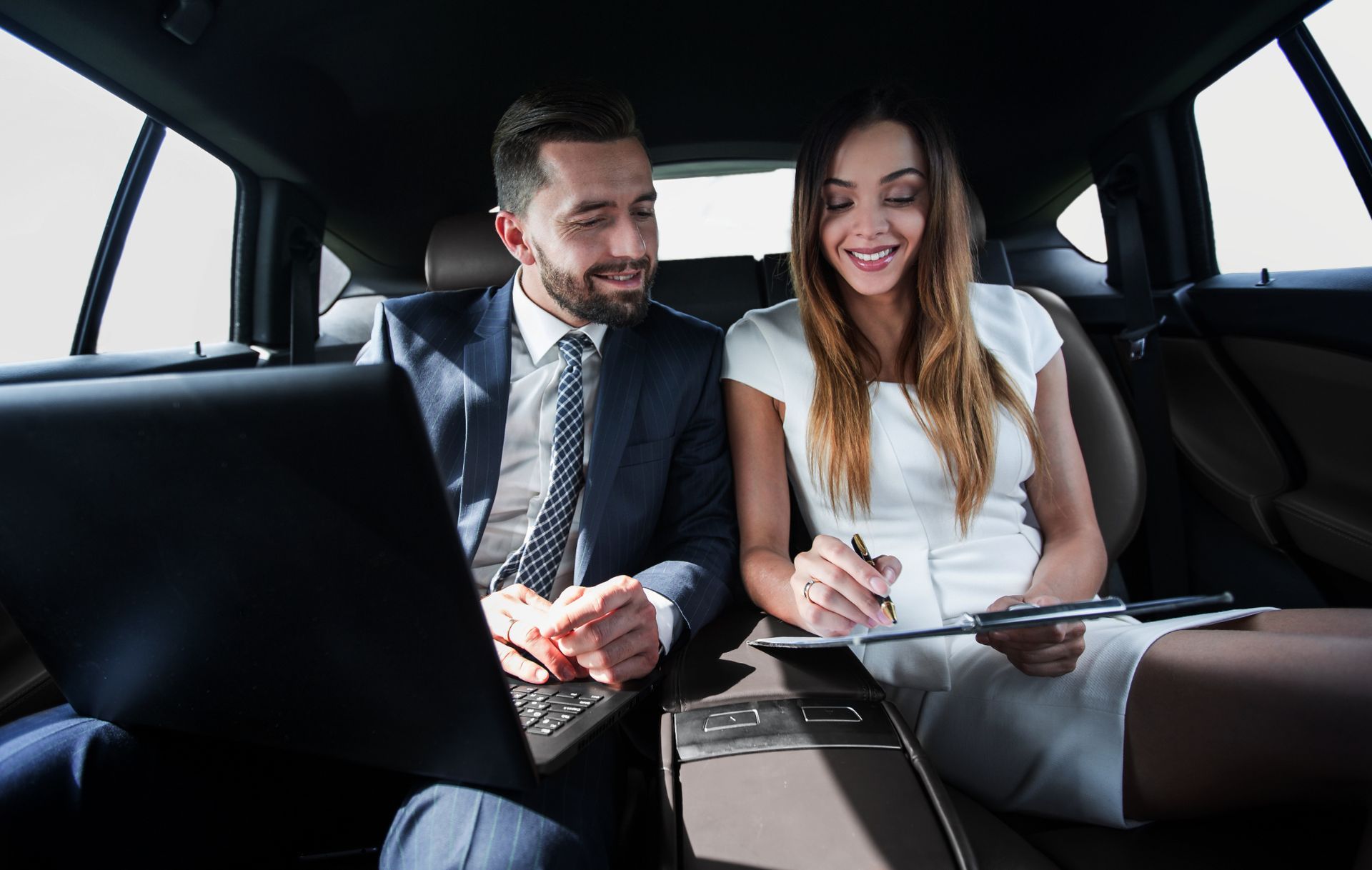 A man and a woman are sitting in the back seat of a car looking at a laptop.