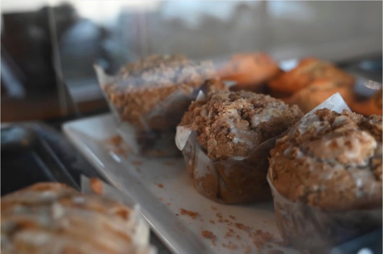 A close-up of several textured, brown muffins sitting on a white tray in a bakery display case.