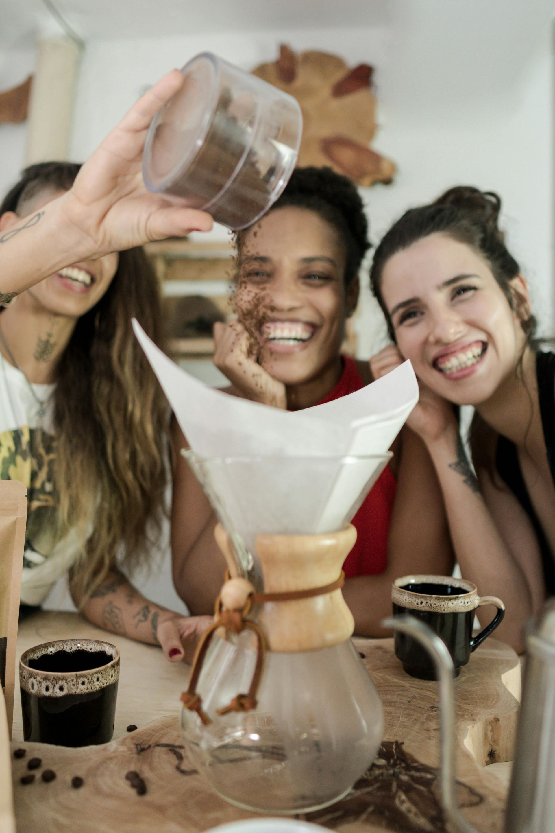 Three people laughing as coffee grounds are poured over a pour-over coffee maker.