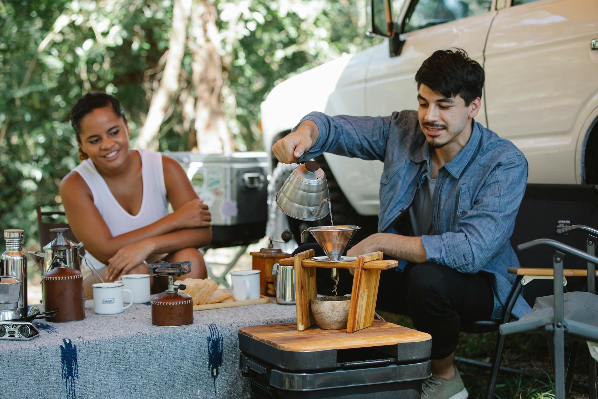 A person pours water over coffee grounds at a campsite, watched by a person seated nearby.