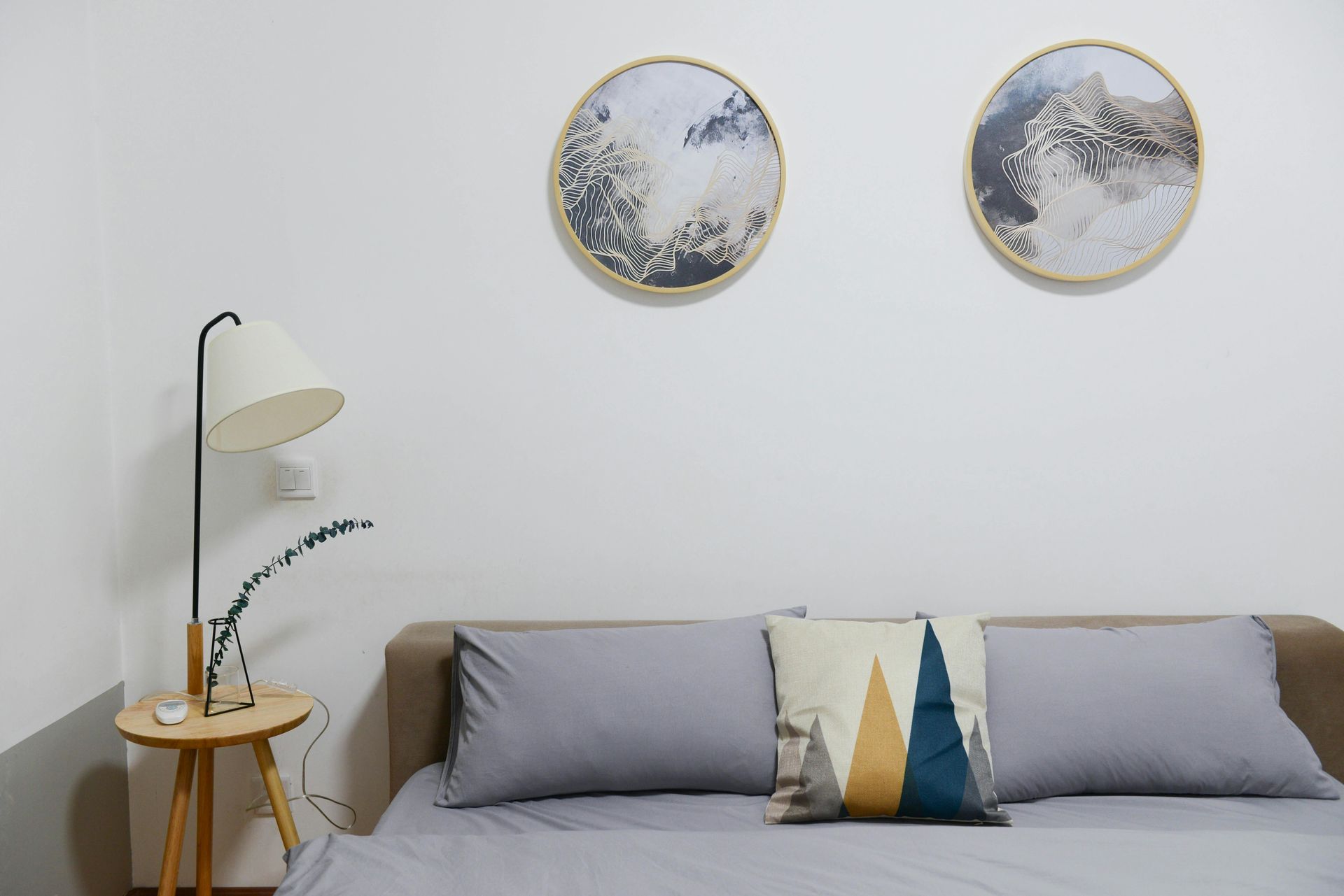 Bedroom with bed, pillows, artwork, lamp, and small table against a white wall.
