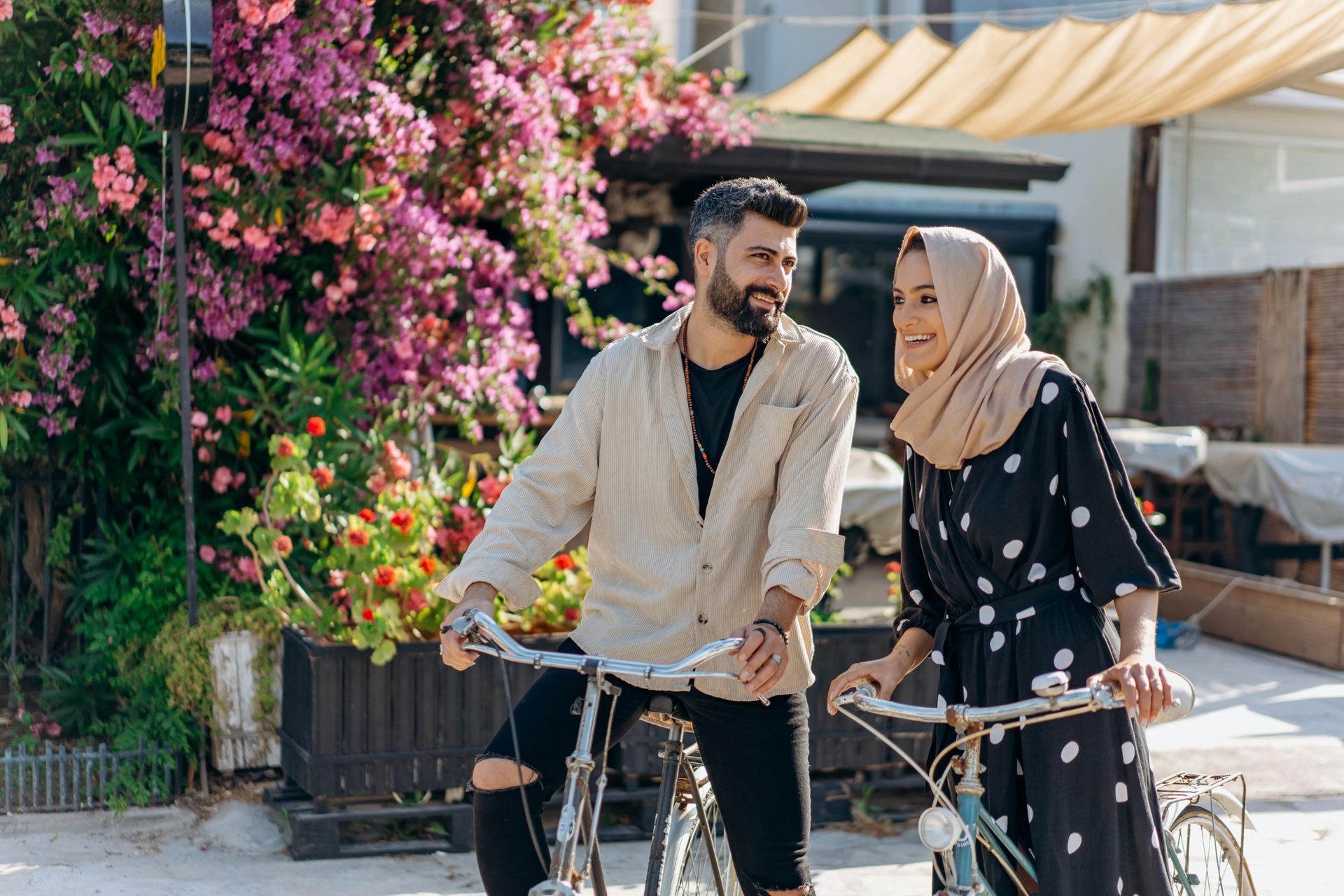 Couple on bikes smiling at each other, near blooming pink flowers and a building.
