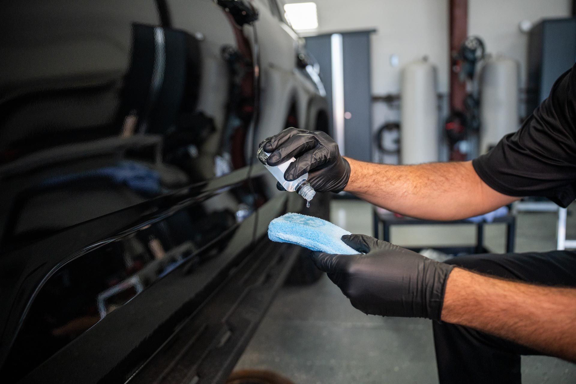 A man wearing black gloves is cleaning a black car with a blue cloth.