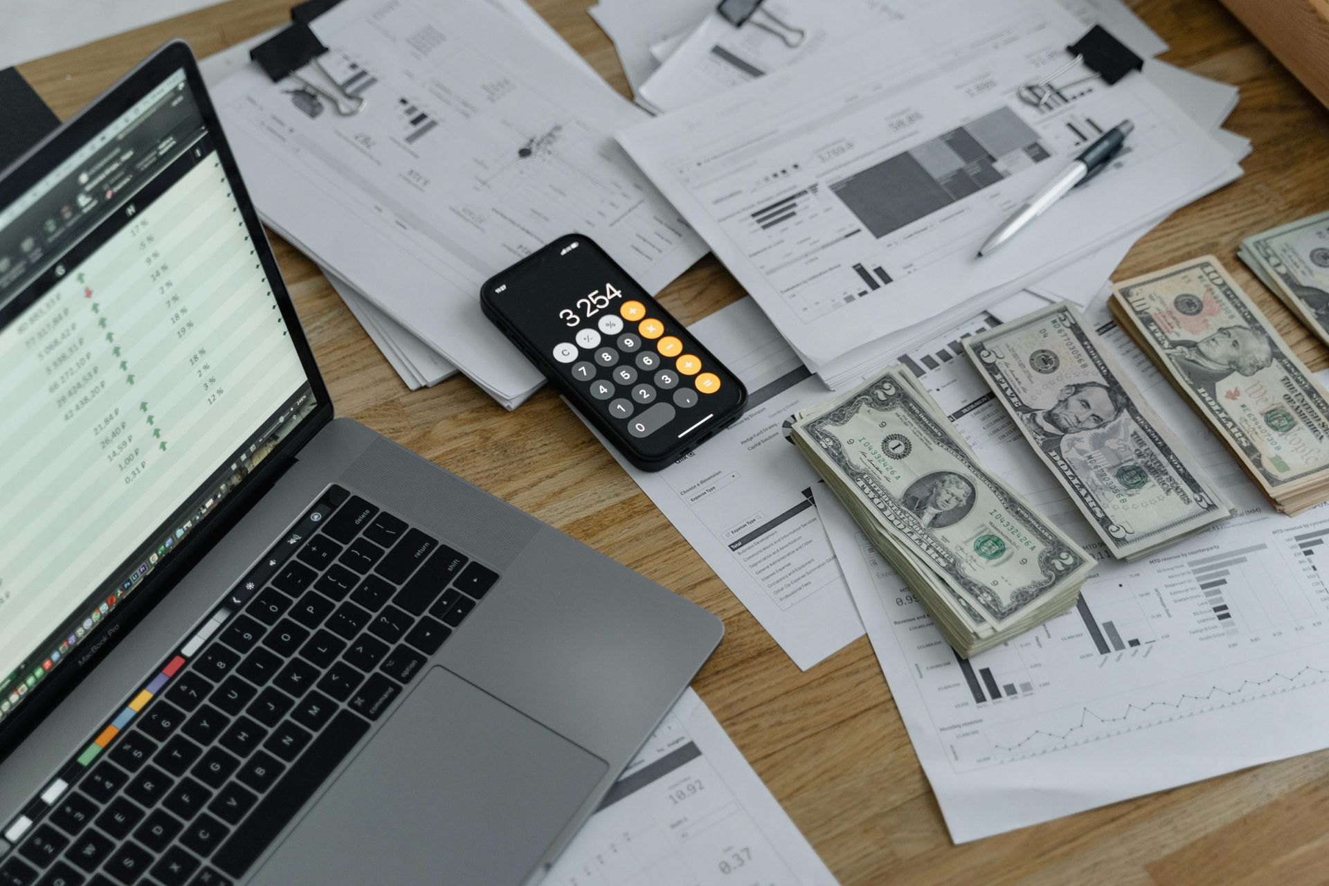 A laptop computer is sitting on a wooden table next to a pile of money and a calculator.