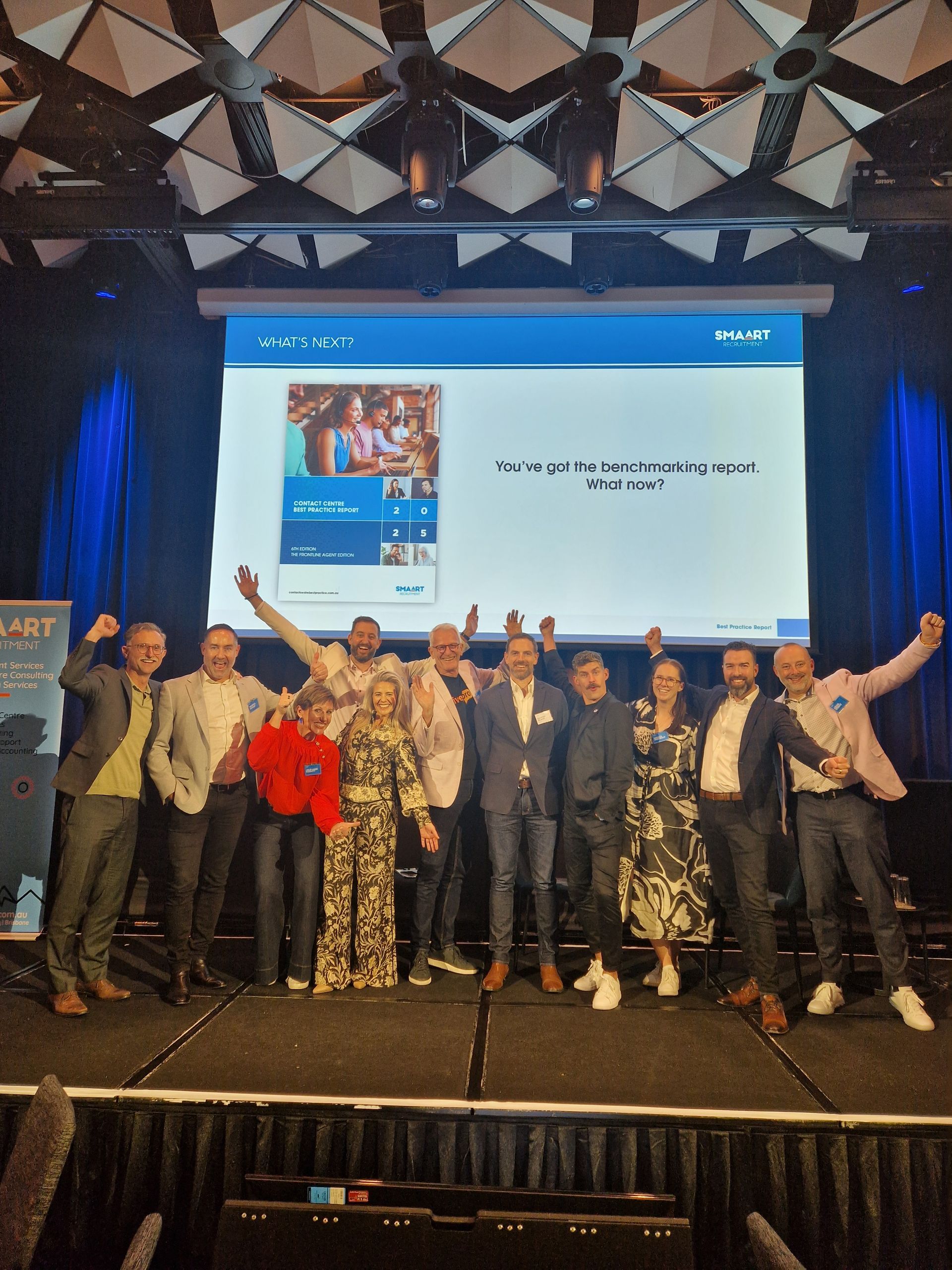 Group of people on stage, posing. Screen behind them, arms raised, celebratory expressions.
