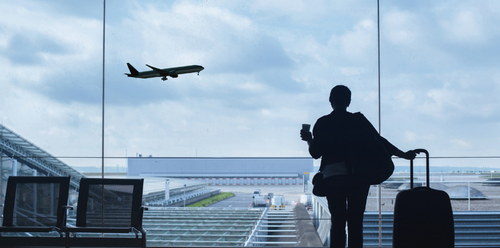Silhouette of a person at an airport window watches a plane take off. Holding a phone with luggage.