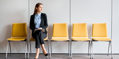Woman in business attire sitting on yellow chair, looking away, waiting room setting.