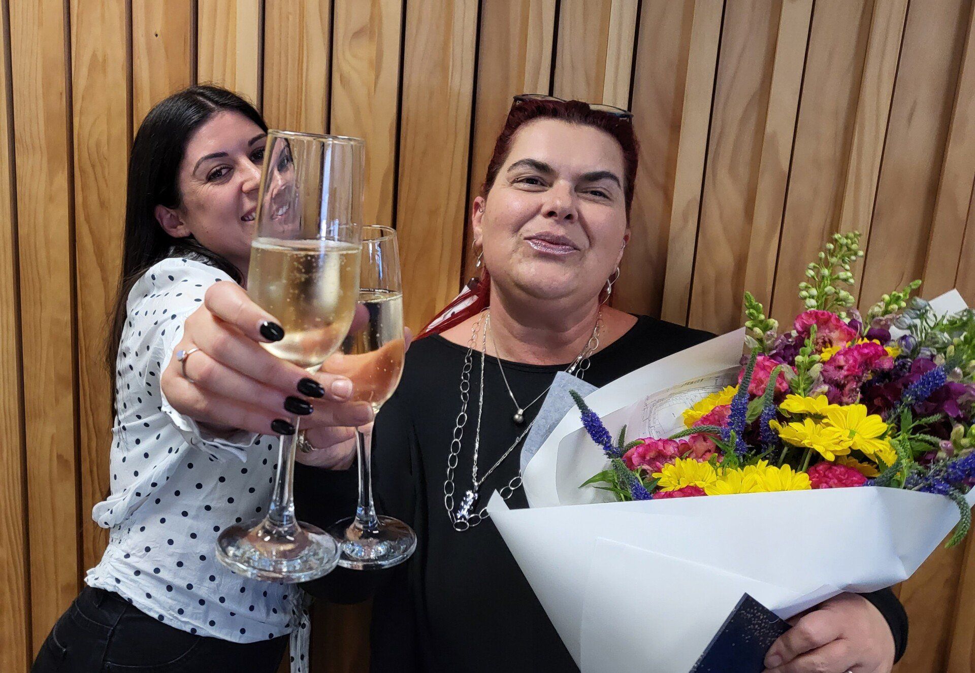 Two women are holding glasses of champagne and a bouquet of flowers.