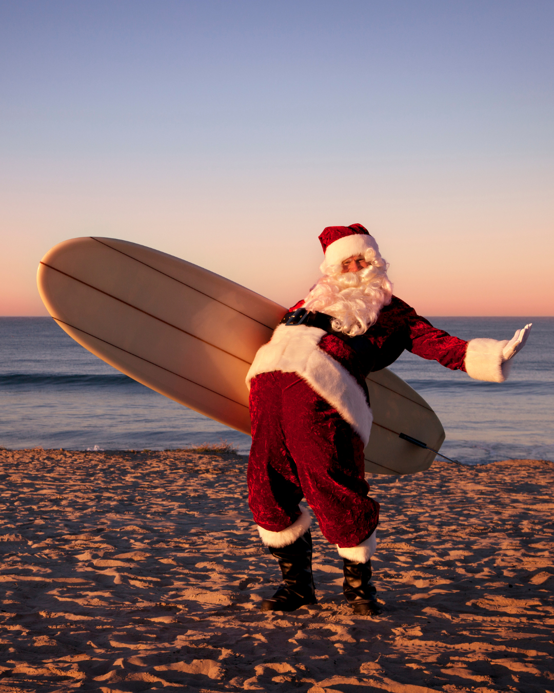 Santa Claus on a beach, holding a surfboard; sun setting, with a wave in the background.