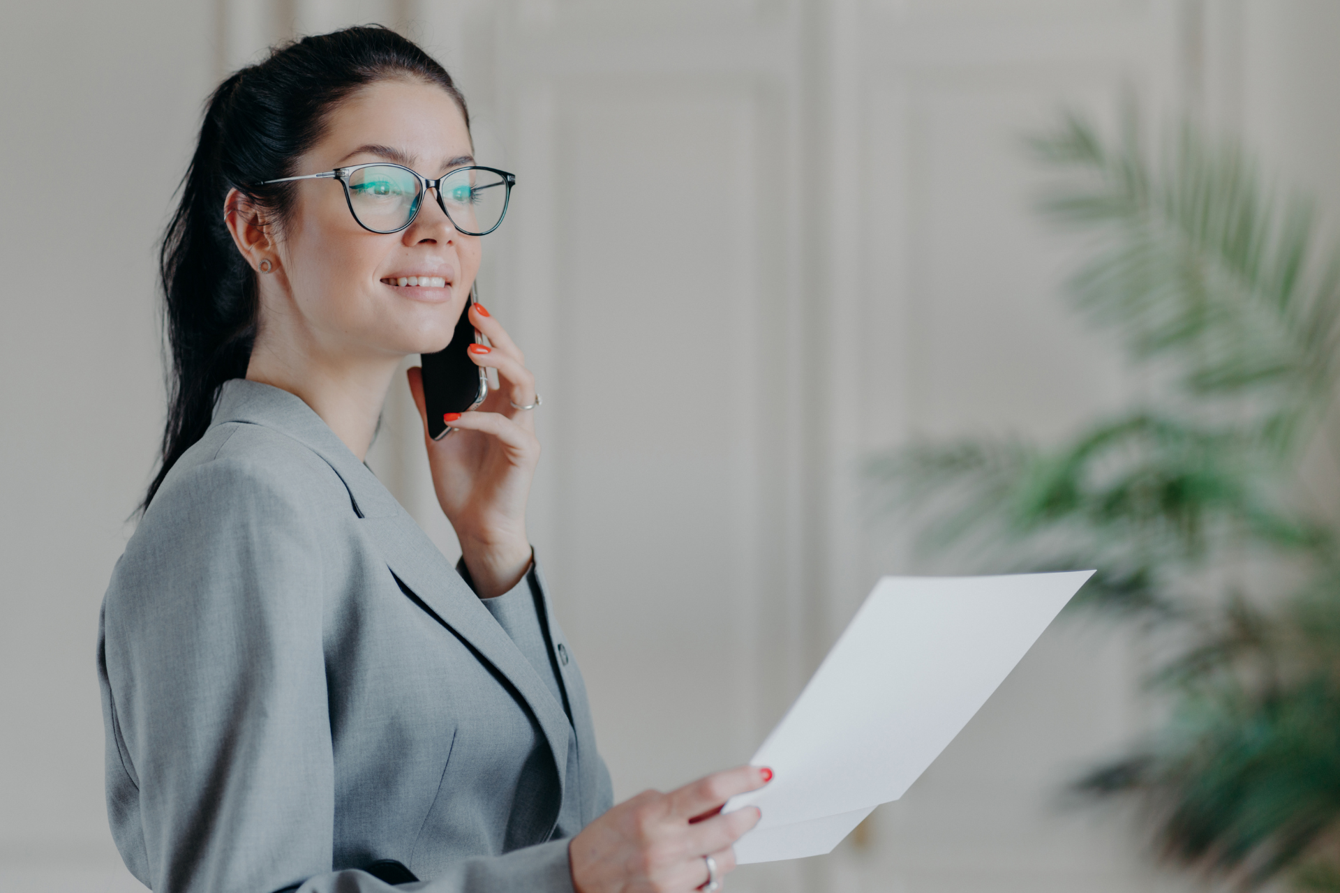 Woman in gray suit talking on phone, holding paper, smiling.