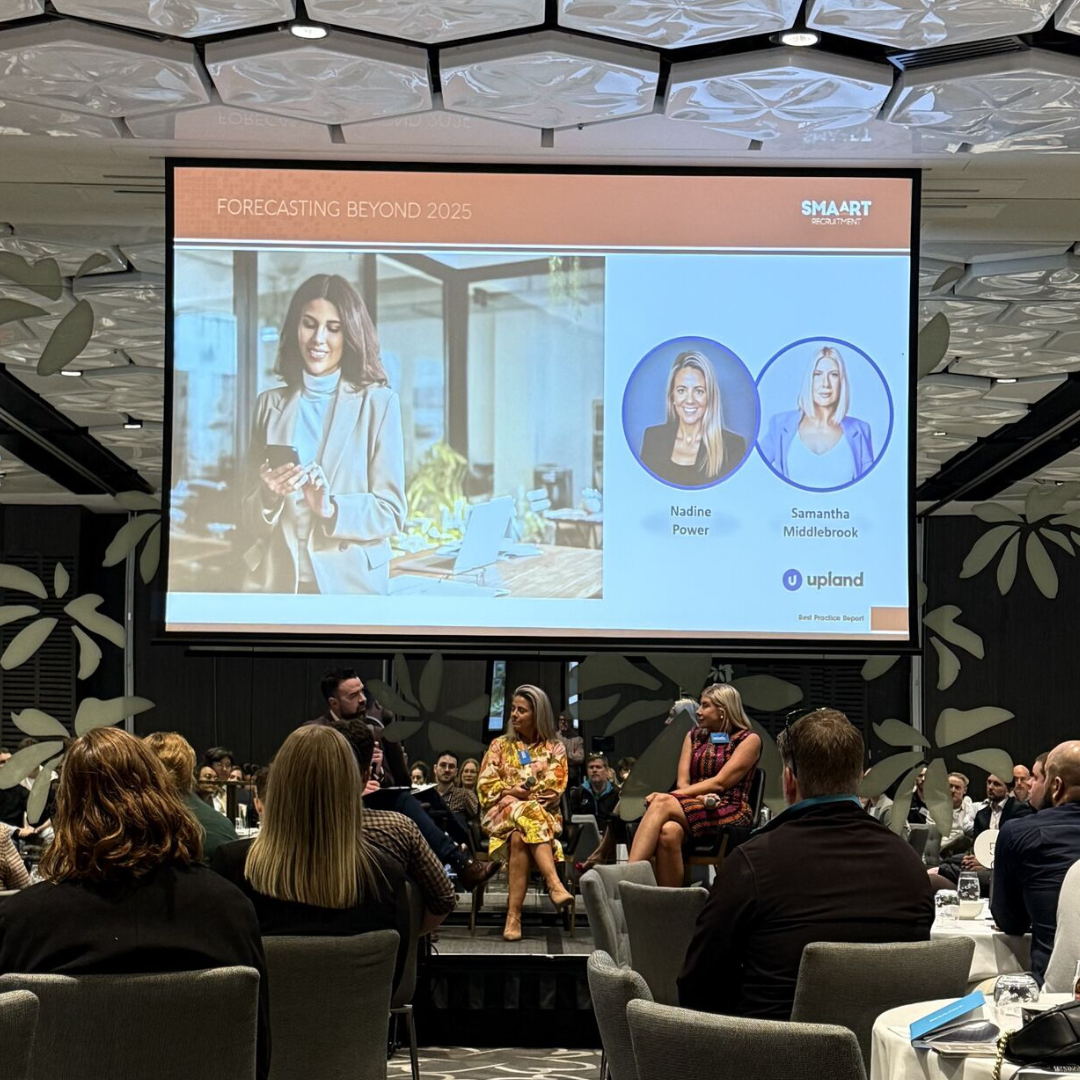 Panel discussion at a conference with speakers on stage, large screen behind them showing presentation