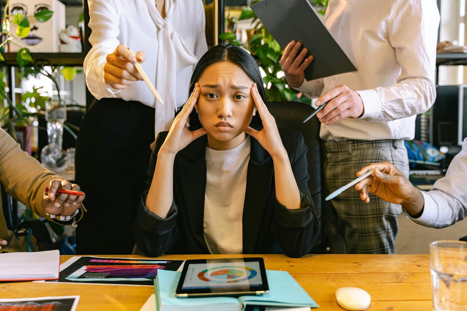 Woman stressed at a desk, surrounded by people pointing and holding documents in an office setting.
