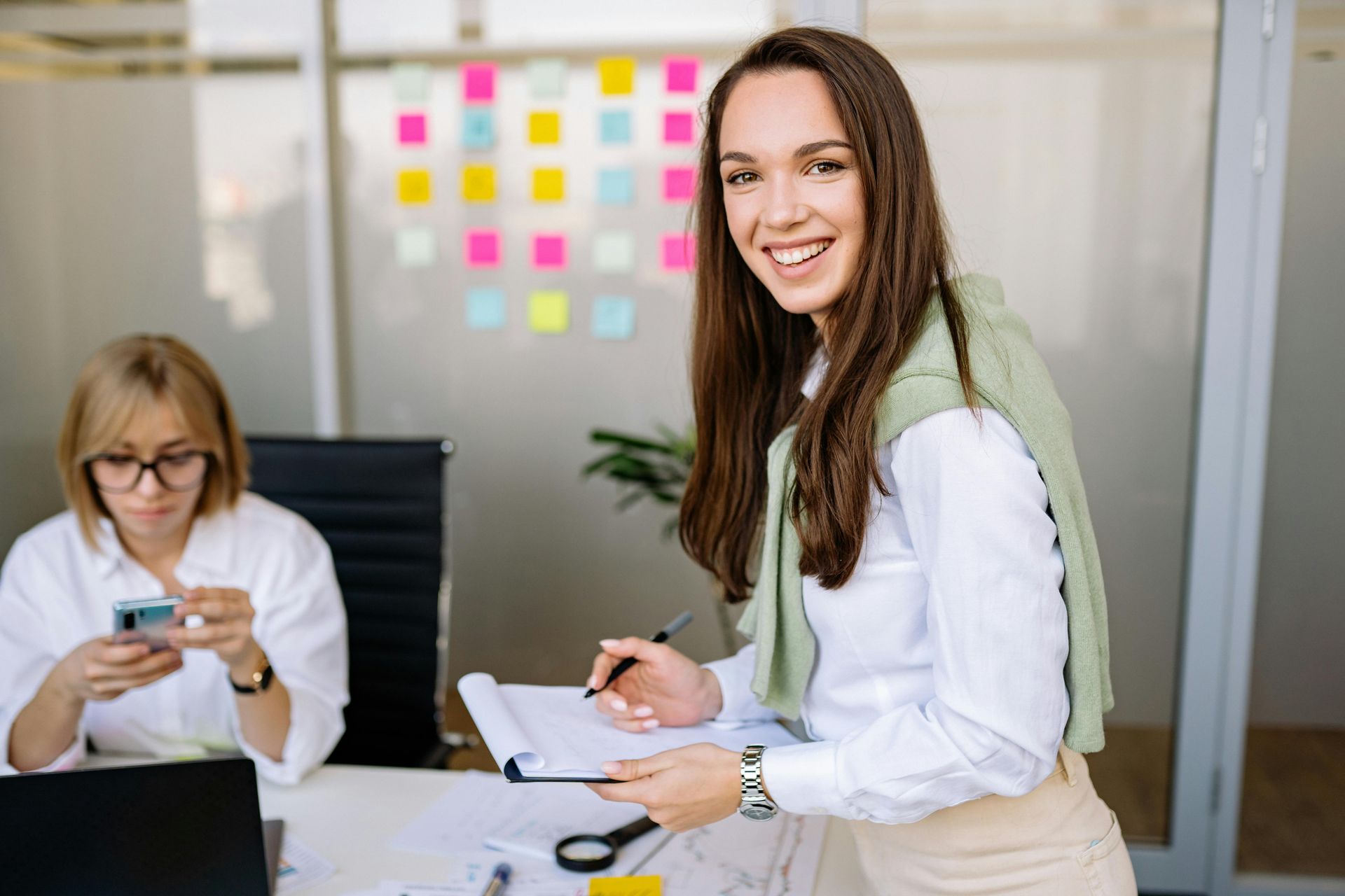 Woman smiling in an office, holding a notebook and pen, with a coworker using a phone in the background.