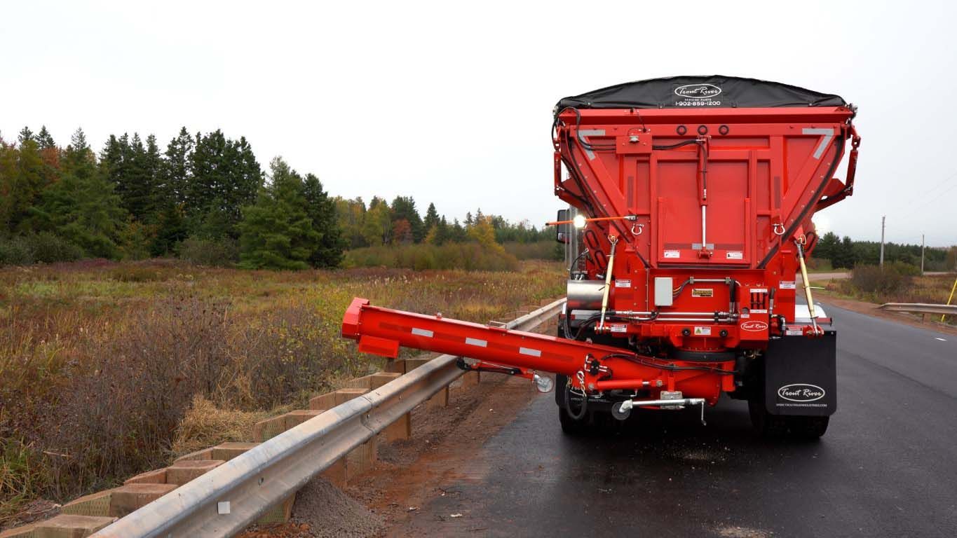Trout River Industries truck-mounted live bottom auger dispensing gravel over a bridge guard rail view from the back