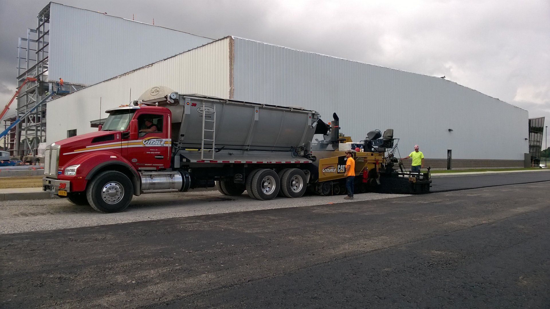 Trout River Industries Truck mounted live bottom unloading asphalt into a paver while a crew pave a road