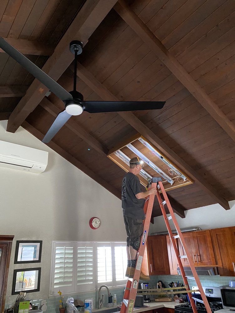 A man is standing on a ladder fixing a ceiling fan in a kitchen.