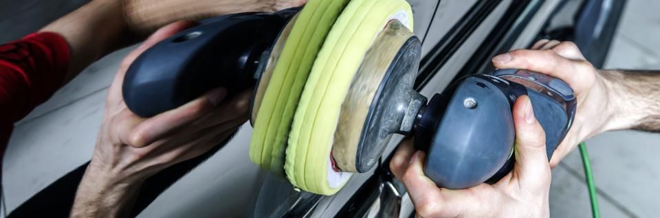 Hands holding two car polishers on a car panel, one with a yellow pad.