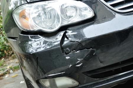 Damaged black car bumper with cracks and scrapes near the headlight.