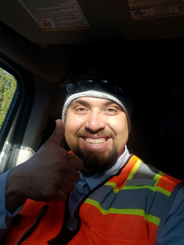 Man in orange vest and beanie gives thumbs up, smiling brightly in a sunny car.