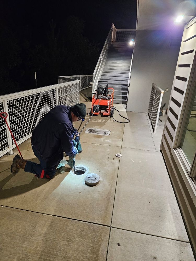 Person kneeling, using a drill on concrete with two open manholes. Staircase and equipment visible at night.