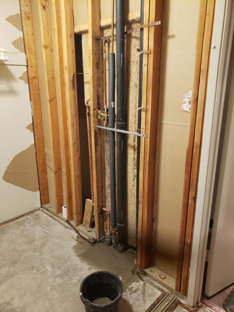 Interior view of exposed plumbing pipes within a wall. A black bucket sits on a concrete floor.