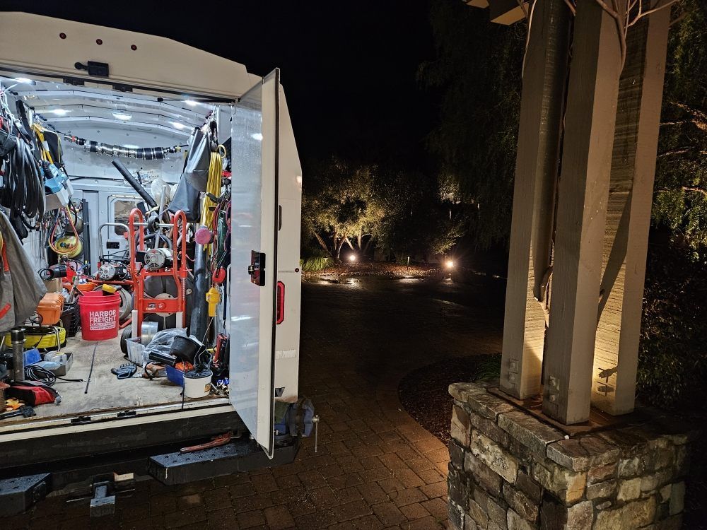 A well-lit work van with open doors, tools inside, and a landscape with a lighted sculpture at night.