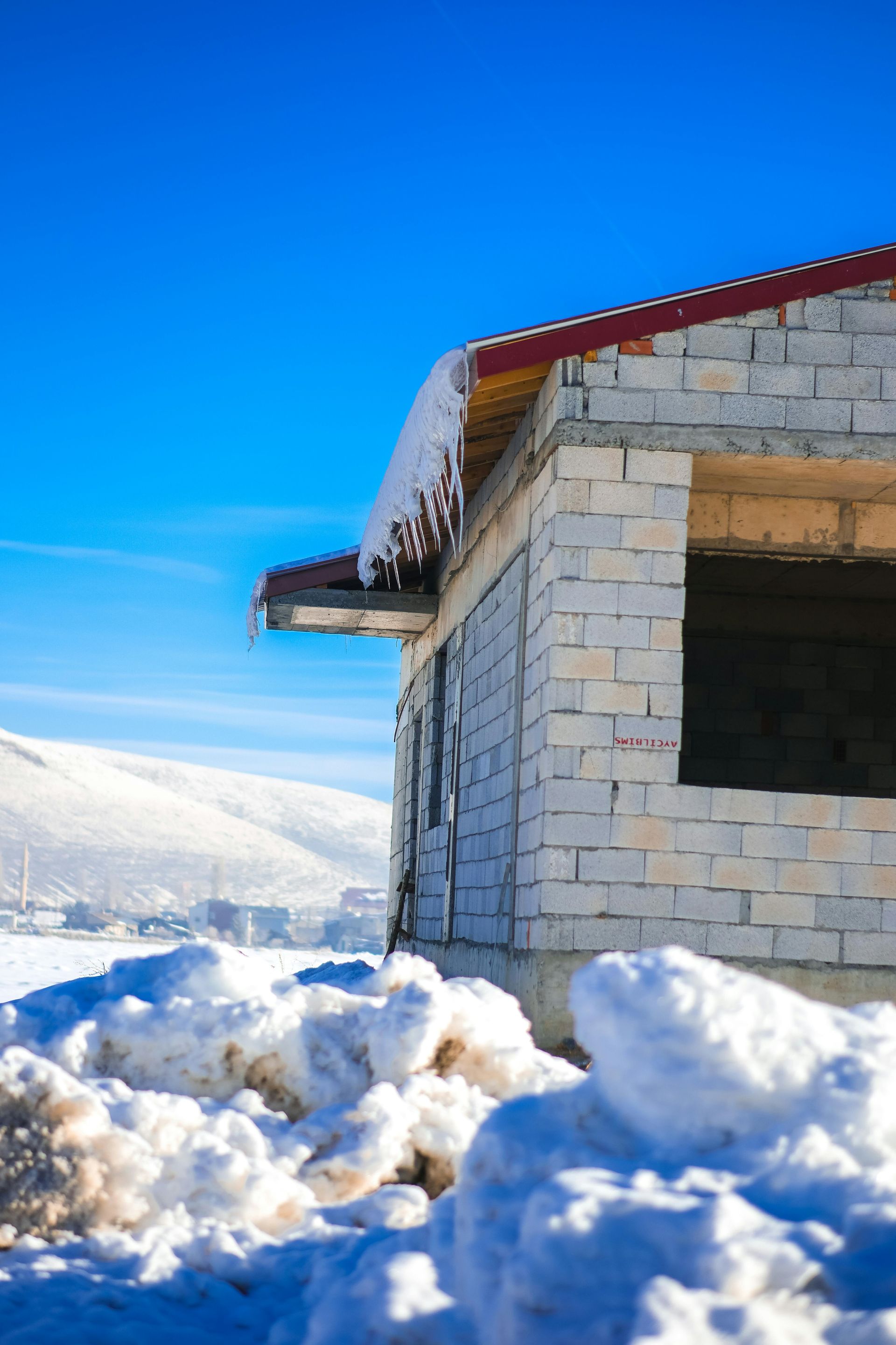 Unfinished brick building with red roof, snow, and ice against a bright blue sky.