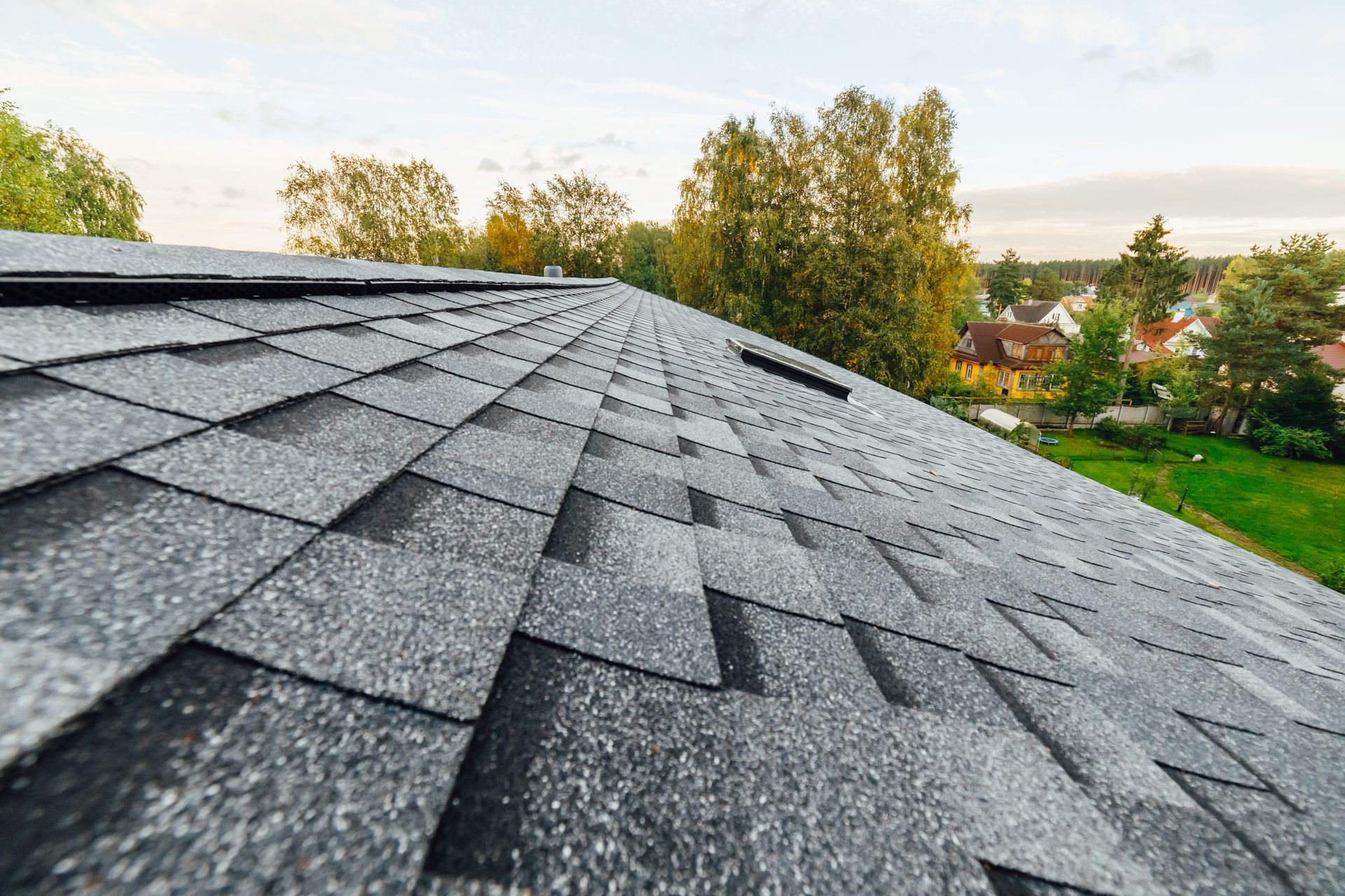 Gray asphalt shingle roof on a house, with trees and a few houses in the background.