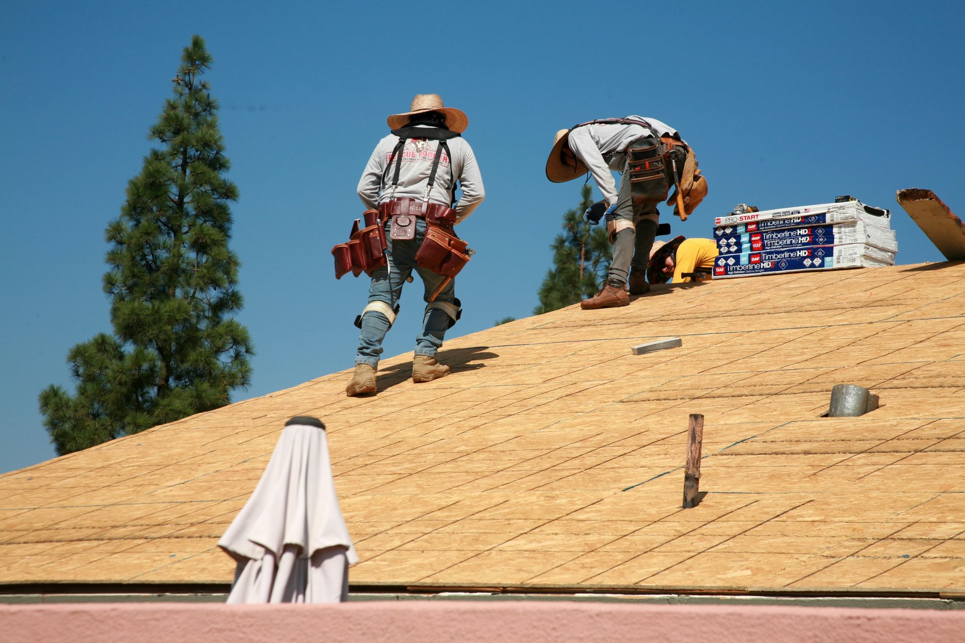 Two roofers working on a sloped roof under a blue sky, wearing safety harnesses and tool belts.