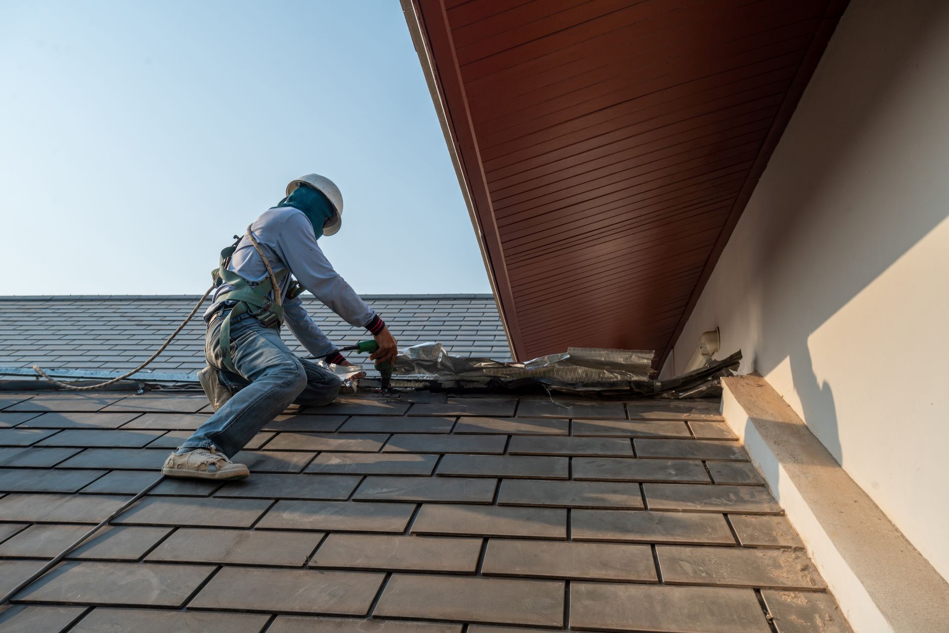 Roofer in safety gear repairs shingles on a roof under a blue sky, near the edge of a house.