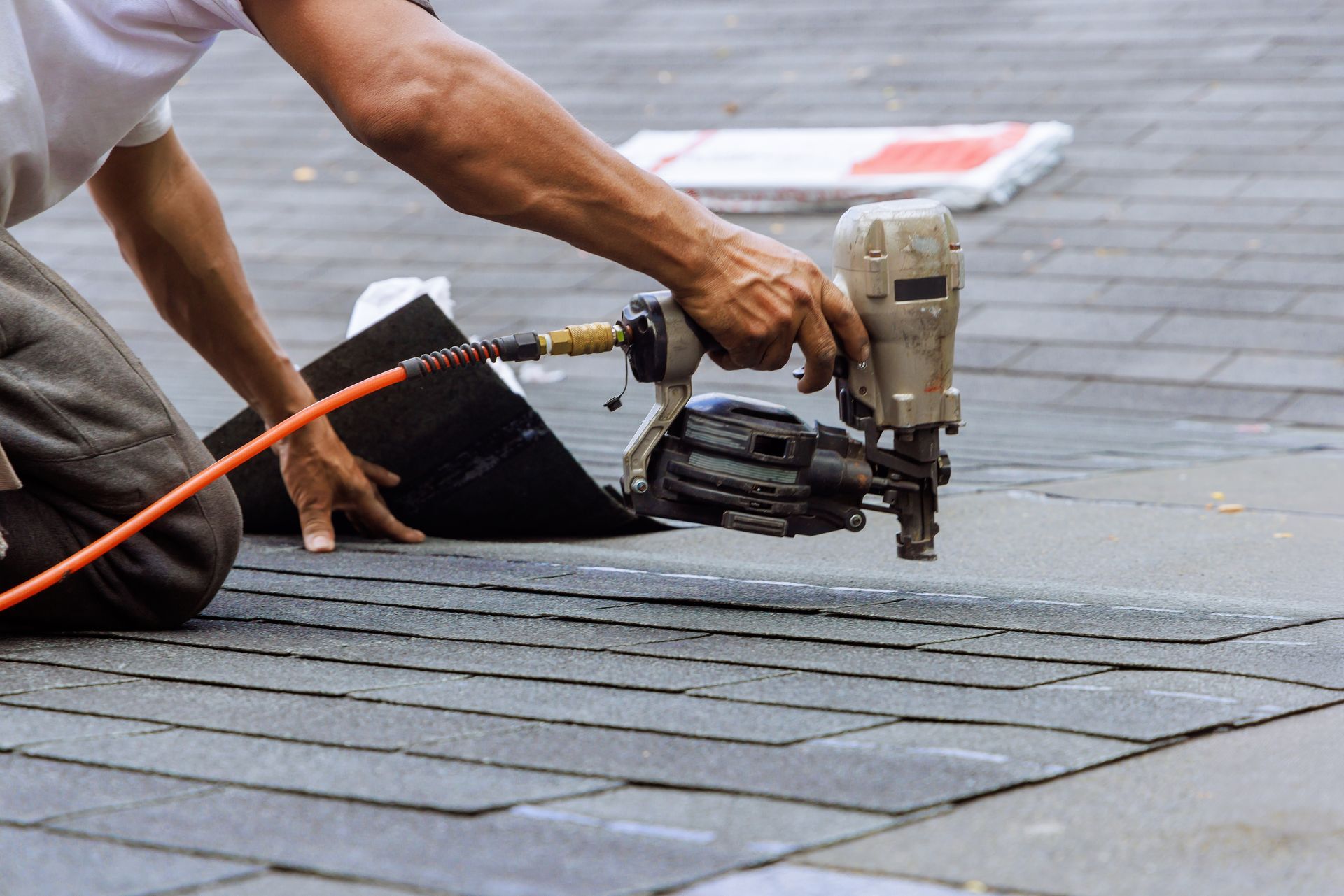 Roofer using a nail gun to attach asphalt shingles to a dark gray roof.