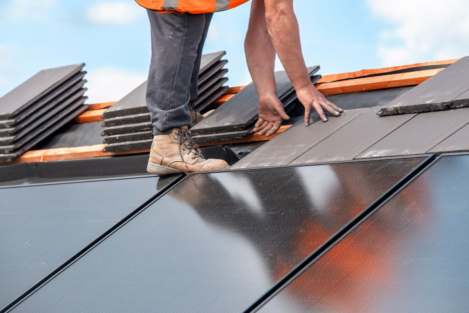 Roofer installing dark gray tiles on a roof with solar panels, under a blue sky.