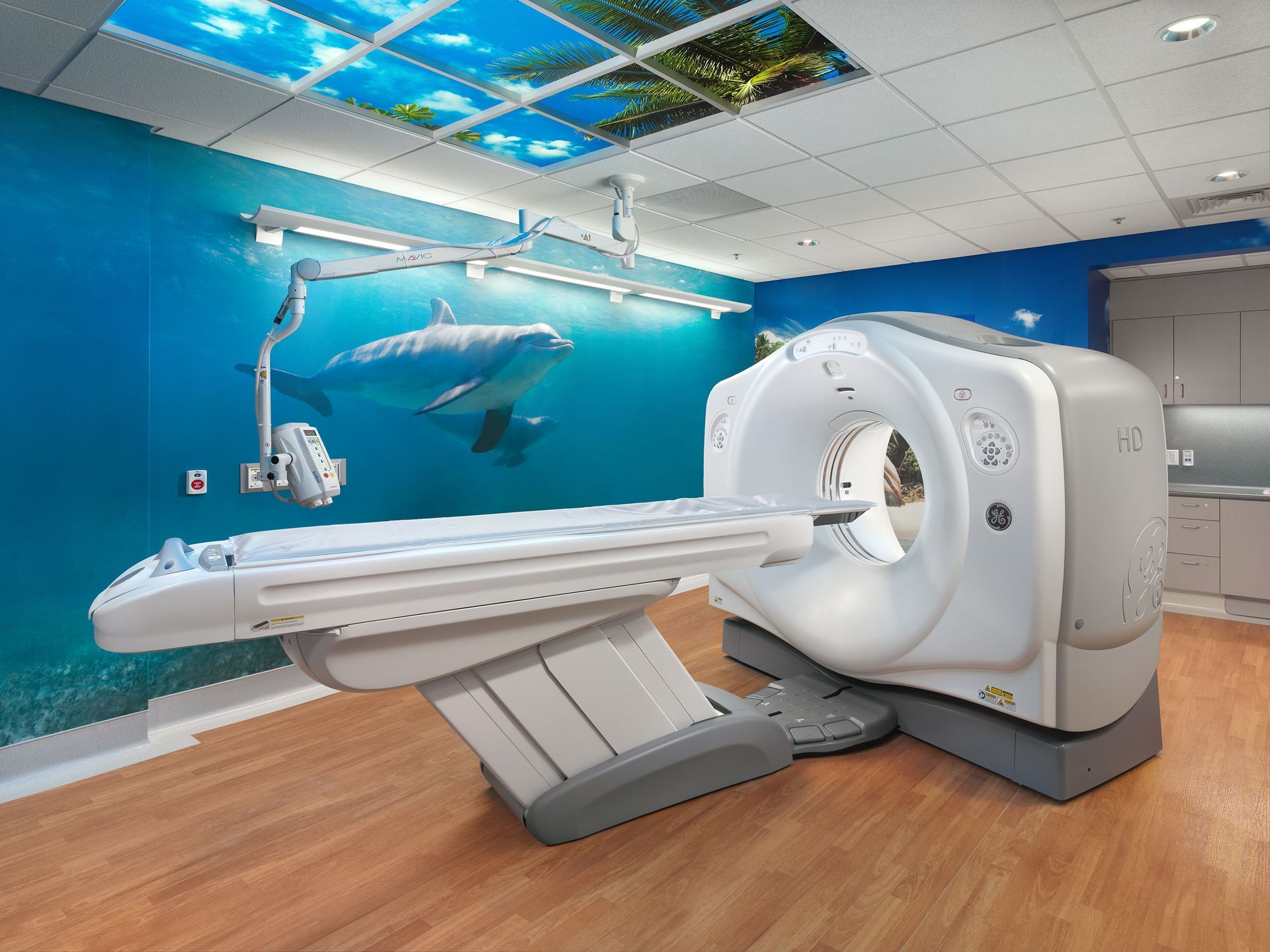 A pediatric CT scanner in a room decorated with underwater-themed wall murals, dolphin imagery, and a sky-view ceiling.