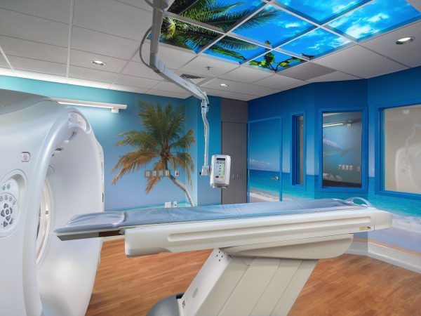 A medical CT scan room decorated with a tropical palm tree mural and a blue sky ceiling panel for a calming atmosphere.