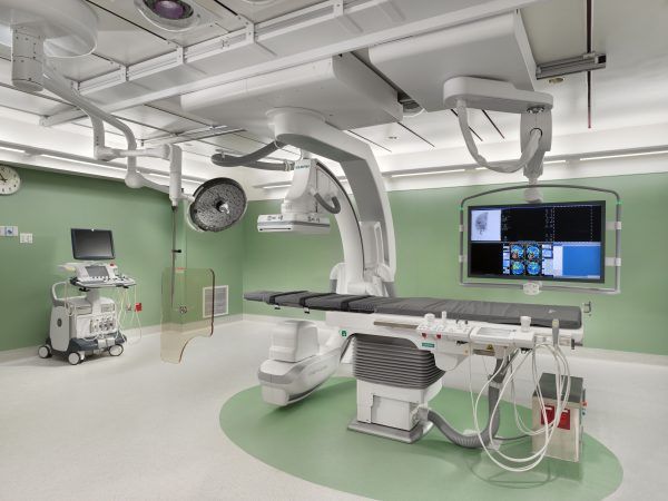 A medical interventional suite featuring a C-arm imaging system, an operating table, and large diagnostic monitors.