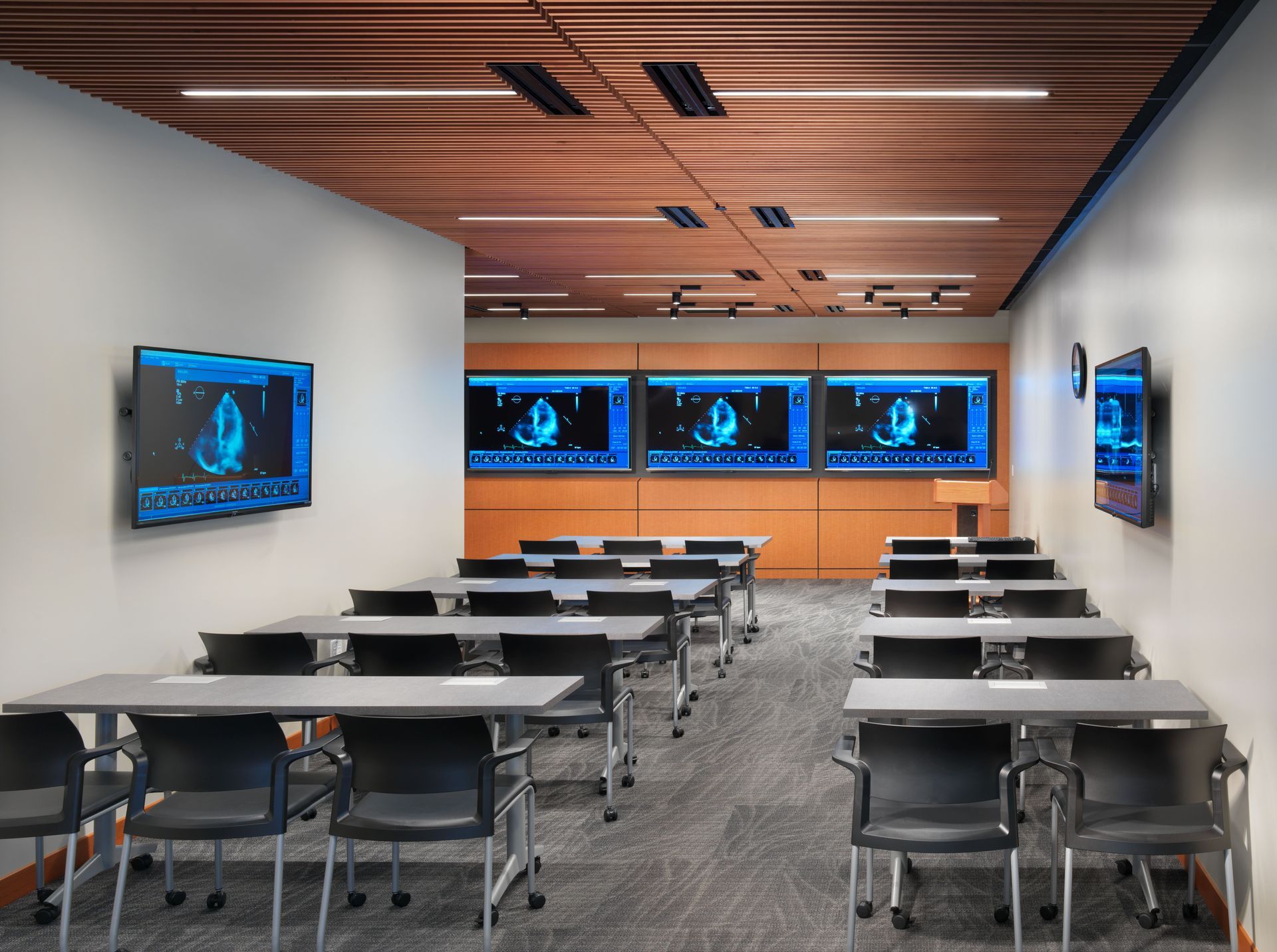 A modern conference room with rows of tables and chairs, wood-slat ceiling, and screens displaying medical imaging.