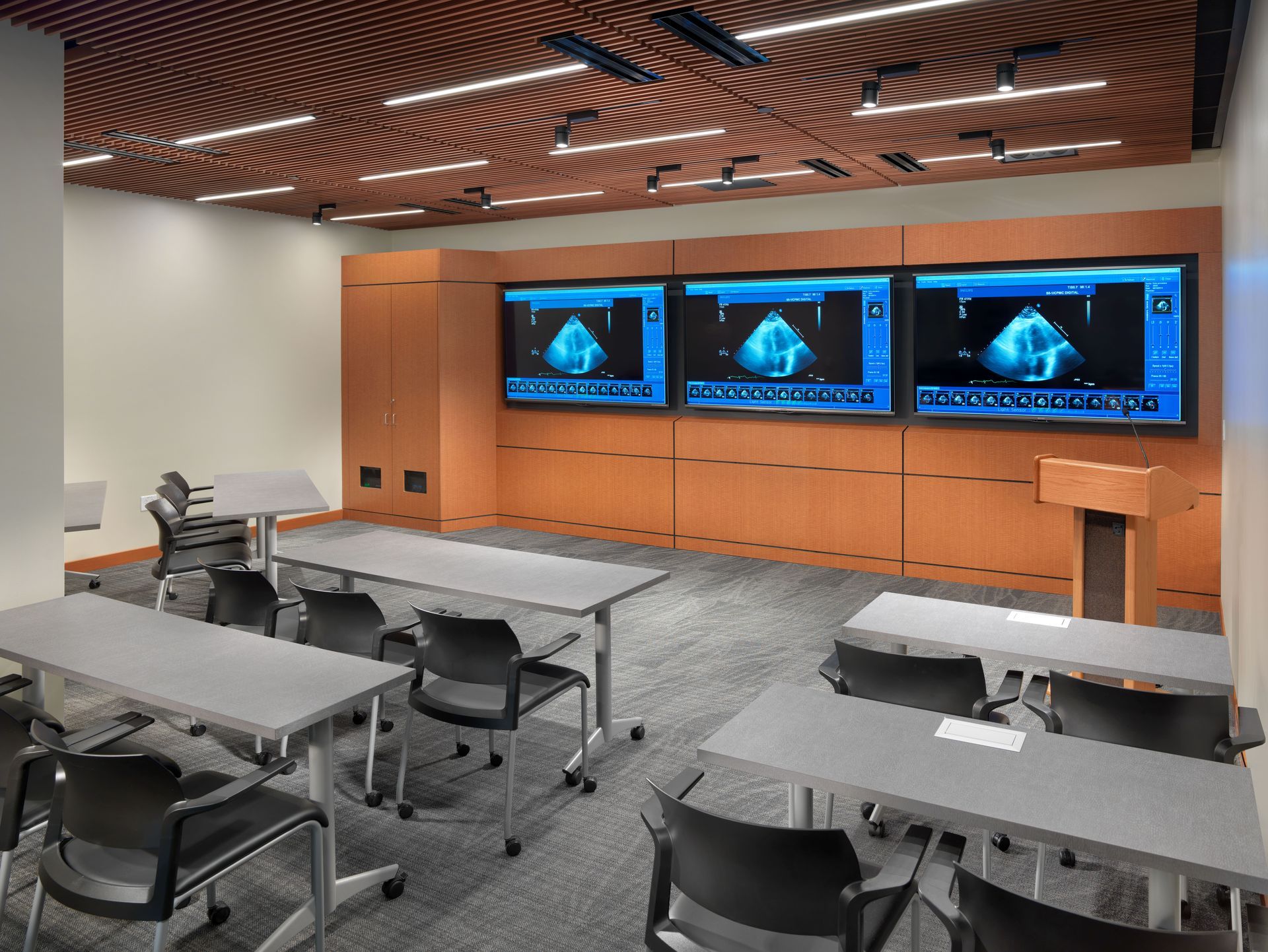 A modern conference room with three large monitors displaying medical ultrasound images above a wooden panel wall.