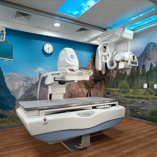A medical fluoroscopy system with a patient table and overhead X-ray tube in a room with a mural of a mountain landscape.