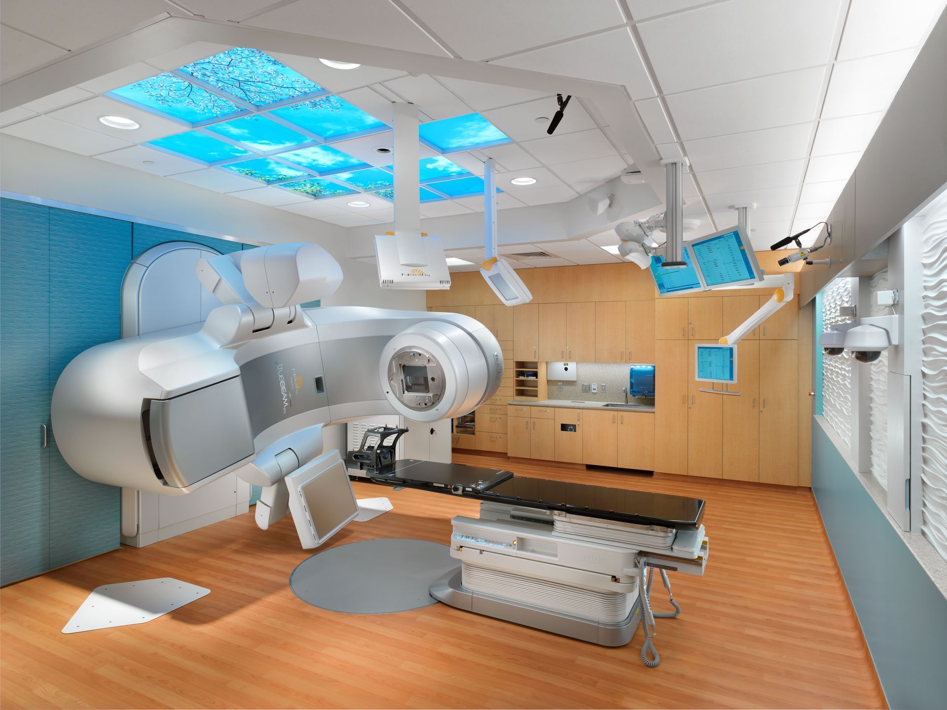 Modern radiation therapy room with a large medical machine, treatment table, and a bright blue ceiling panel display.