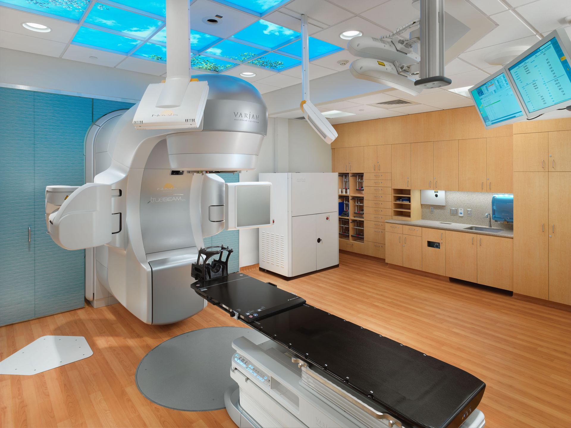A medical linear accelerator for radiation therapy in a treatment room with wood cabinetry and light-blue ceiling panels.