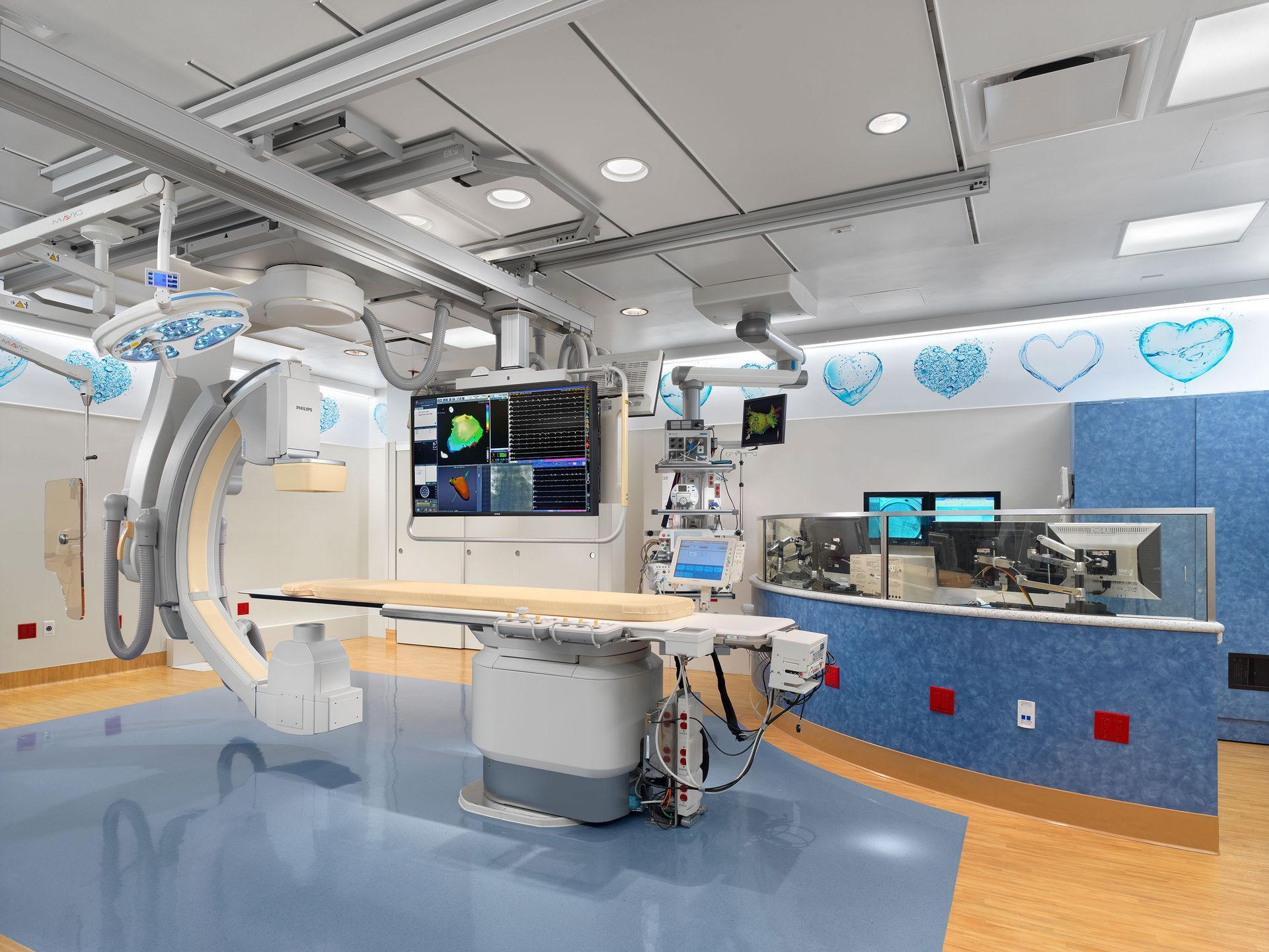 A modern, clean medical catheterization lab featuring an X-ray imaging system, exam table, and blue-toned walls.