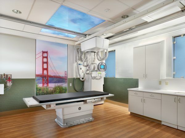 An X-ray imaging room featuring a central exam table, a ceiling-mounted machine, and a wall mural of the Golden Gate Bridge.
